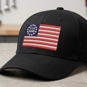 May include: Black baseball cap featuring an embroidered American flag design. The flag includes red and white stripes, a blue field with white stars, and the text "1776 2026".