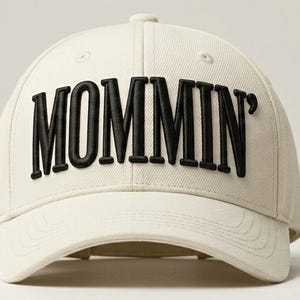 May include: A cream-colored baseball cap featuring the word MOMMIN' in bold, raised black lettering across the front panels. The cap has a structured crown, a curved brim with stitched detailing, and a button at the top center of the dome.