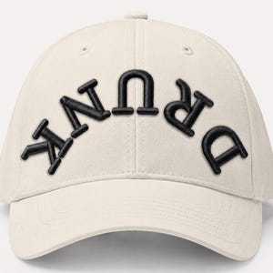 May include: Cream-colored baseball cap with the word "DRUNK" embroidered in black, curved across the front. The hat has a curved brim and a button on top. A casual accessory.