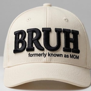 May include: A cream-coloured baseball cap featuring the word "BRUH" in bold, black letters. Beneath, the phrase "formerly known as MOM" is printed in a smaller, black font. The cap has a curved peak.