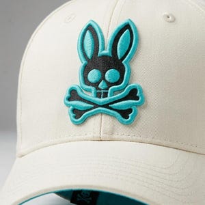 Puede incluir: Gorra de béisbol color crema con un logotipo de calavera de conejo negro y turquesa. La gorra tiene una visera curva y un botón en la parte superior. El logotipo es un parche bordado con una cabeza de conejo y huesos cruzados.