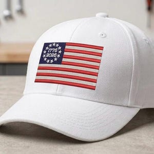 Puede incluir: Gorra de béisbol blanca con un diseño de bandera bordada. La bandera tiene rayas rojas y blancas, un cuadrado azul con estrellas blancas y los números "1776" y "2026". La gorra tiene una visera curva.