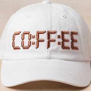 May include: A white baseball cap featuring the word CO:FFEE embroidered in a brown, digital-style font across the front panel. The cap has a structured crown, a curved brim with visible stitching, and a button at the top center of the dome.