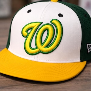 Op de afbeelding: Een honkbalpet met een witte voorkant en een gele klep. De pet heeft een groot, gestileerd geel en groen "W" logo. De achterkant en zijkanten zijn groen en de bovenste knop is geel.