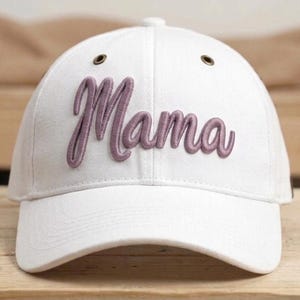 Könnte beinhalten: Eine weiße Baseballkappe mit dem Wort Mama, das in einer erhabenen, malvenfarbenen Schreibschrift auf die Vorderseite gestickt ist. Die Kappe verfügt über einen gebogenen Schirm, einen Knopf oben und metallische Ösen zur Belüftung am Oberkopf.