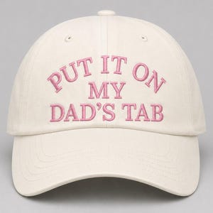 May include: An off-white baseball cap featuring the embroidered text "PUT IT ON MY DAD'S TAB" in pink thread. The cap has a structured crown, a curved brim, and stitched eyelets on the top panels, presenting a clean and simple design.