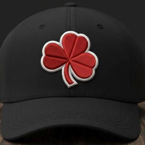 May include: Black baseball cap featuring a red and white embroidered shamrock. The shamrock, a three-leaf clover, is a symbol of luck and Irish heritage. The cap has a curved peak and a structured fit.