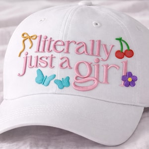 Puede incluir: Gorra de béisbol blanca con la frase bordada "literally just a girl!" en rosa. El diseño incluye un lazo amarillo, dos mariposas azules, cerezas rojas y una flor morada.