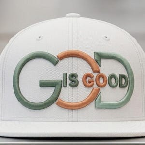 Op de afbeelding: Een witte baseballpet met verhoogde, geborduurde letters op de voorkant. Het ontwerp toont de tekst GOD IS GOOD, waarbij de letters G en D in een gedempt saliegroen zijn en de letters O en IS GOOD in een metaalachtige koperkleurige draad.
