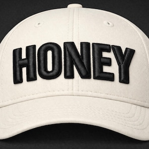 May include: Cream-colored baseball cap with the word "HONEY" in bold, black, raised lettering across the front. The cap has a curved brim and a button on top. The background is dark.