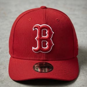 Può includere: Un cappellino da baseball rosso a tinta unita con una lettera B in rilievo ricamata sul pannello frontale, delineata in bianco. Il cappellino presenta un adesivo circolare 59FIFTY sulla visiera piatta, con il testo 59FIFTY e una taglia.