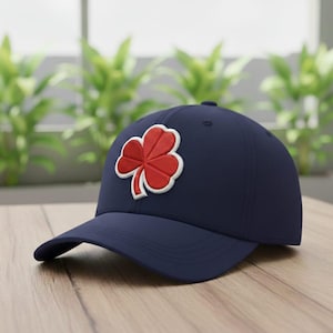 May include: A navy blue baseball cap featuring a red and white embroidered shamrock on the front. The cap has a curved brim and a structured crown. The hat is sitting on a wooden surface.