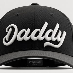 May include: A black baseball cap featuring the word Daddy embroidered in white, raised script lettering across the front panels. The cap has a structured crown, a curved brim, and a small interior label that reads Premium Caps in white text.