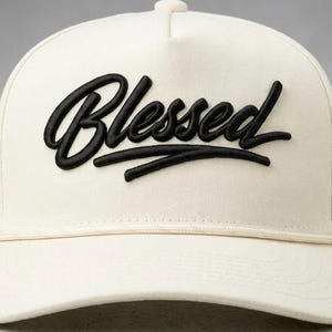 May include: A cream-coloured baseball cap featuring the word "Blessed" embroidered in black script. The cap has a curved peak and a structured crown. The design is simple and modern, suitable for everyday wear.