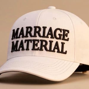 Könnte beinhalten: Weisse Baseballkappe mit dem Schriftzug "MARRIAGE MATERIAL" in fetten, schwarzen Buchstaben. Die Kappe hat einen gebogenen Schirm und einen Knopf oben. Klassischer Stil.