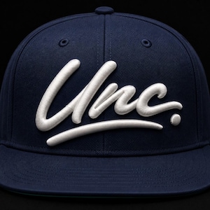 May include: A navy blue baseball cap featuring the word Unc. embroidered in raised white lettering across the front panels. The cap has a structured crown, a flat brim, and a button on top, showcasing a clean and bold design with high-contrast colors.