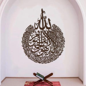 May include: A brown, circular Islamic calligraphy wall art piece. The artwork features intricate Arabic script, mounted on a white wall. A wooden book stand with a green book and a string of beads are in the foreground.