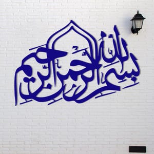 May include: A blue Arabic calligraphy wall art piece against a white brick wall. The artwork features elegant script in a stylized design. A black outdoor lamp is mounted on the wall to the right of the artwork.