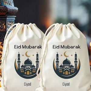 May include: Two cream-coloured drawstring bags, each printed with "Eid Mubarak" and a navy and gold mosque design. The bags also feature the word "Ciyad". They have white drawstrings.