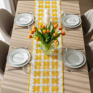 May include: A dining table set for a meal, featuring a yellow and white crochet table runner. The table is set with white plates, silverware, and clear glasses. A vase of orange and yellow tulips and two white candles are centered on the table.