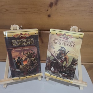 以下が含まれることがあります： リチャード・A・ナーク著のドラゴンランス・ファンタジー小説『The Minotaur Wars Volume One Night of Blood』と『Volume Two Tides of Blood』の2冊。2つの小さな木製イーゼルに飾られており、各表紙にはミノタウロスの戦士の緻密なイラストが描かれています。