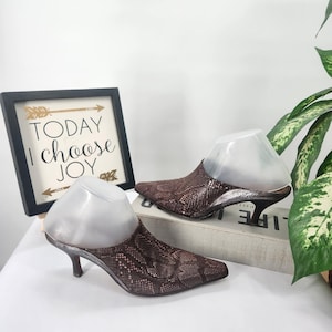 Peut inclure: Une paire de mules marron à motif peau de serpent avec des bouts pointus et des talons aiguilles. Les chaussures présentent un éclat métallique et sont présentées sur des formes en plastique. Un cadre affiche le texte : TODAY I choose JOY.