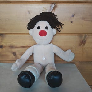 Puede incluir: Una muñeca de peluche hecha a mano con un cuerpo beige, cabello negro y una nariz roja. La muñeca tiene ojos negros de botón, calcetines blancos y zapatos negros. La muñeca está sentada sobre una superficie gris claro.