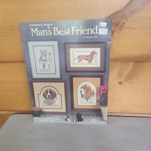 May include: A cross-stitch pattern book titled "Man's Best Friend" with framed dog portraits. The cover shows the title and "Collection Two" with images of a Schnauzer, Dachshund, Springer Spaniel, and Basset Hound. The book is on a gray surface.