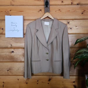 May include: A tan blazer featuring a notched lapel, two front buttons, and two horizontal welt pockets. The blazer has long sleeves with three small buttons at each cuff and is displayed on a wooden hanger against a horizontal wood plank wall.