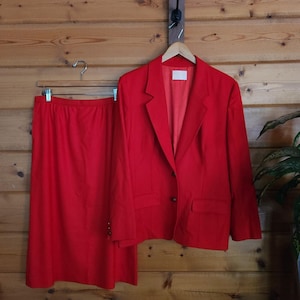 Op de afbeelding: Een felrood rokpak, bestaande uit een blazer en een knielange rok, hangt tegen een houten muur. De blazer heeft een reverskraag, twee voorzakken en een sluiting met twee knopen. De rok is effen, helderrood.