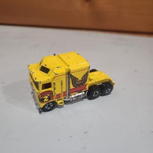 May include: A yellow die-cast toy semi-truck cab featuring a black eagle graphic on the side, a red stripe, and the number 2 printed in yellow on a black circle. The truck has silver-colored exhaust stacks and black wheels with silver rims.