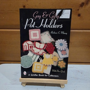 May include: A black book titled Gay & Gifty Pot Holders by Barbara E. Mauzy, featuring a collection of colourful crocheted pot holders in various shapes like flowers, dresses, and squares, with a Schiffer Book for Collectors label at the bottom.