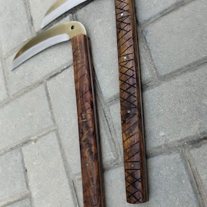 Puede incluir: Dos cuchillas curvas con mangos de madera. Las cuchillas son de color plateado y los mangos son de color marrón oscuro con patrones de trama cruzada y diagonales tallados. Las herramientas probablemente se utilizan para cortar o cosechar.