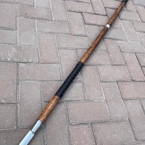 May include: A spear with a wooden shaft and a metal blade. The shaft is brown with a black grip near the center and a silver band. The spear is lying on a brick surface.