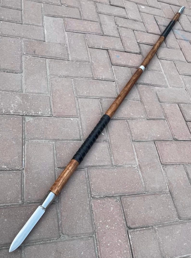 May include: A spear with a wooden shaft and a metal blade. The shaft is brown with a black grip near the center and a silver band. The spear is lying on a brick surface.