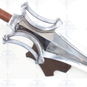 May include: A close-up of a silver sword with a brown leather-wrapped handle. The blade is polished and sharp, with a decorative hilt. The sword is mounted on a wooden stand. The text "Crafted Legends Edge" is visible.