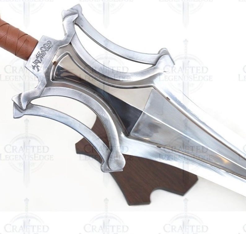 May include: A close-up of a silver sword with a brown leather-wrapped handle. The blade is polished and sharp, with a decorative hilt. The sword is mounted on a wooden stand. The text "Crafted Legends Edge" is visible.