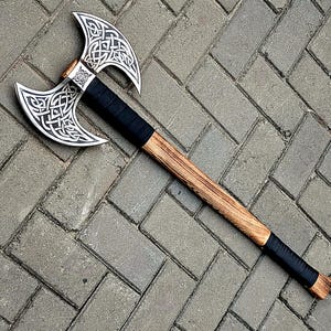 May include: A double-bitted axe featuring a silver-colored metal head with intricate engraved knotwork patterns. The handle is crafted from light brown wood, accented with black wrapped grips located near the head and at the base of the shaft.