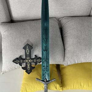 May include: A decorative sword with a teal blade and ornate silver handle. The blade features etched designs. A black cross with gold accents is also present. The items are displayed on a white surface with grey and yellow cushions in the background.