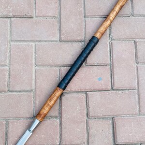 May include: A spear with a wooden shaft and a metal blade. The shaft is light brown with a black grip near the center. The blade is silver and tapers to a sharp point. The spear is lying on a brick surface.