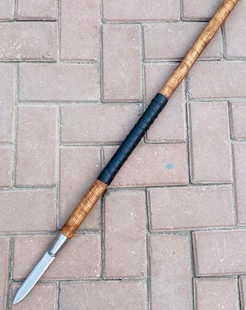 May include: A spear with a wooden shaft and a metal blade. The shaft is light brown with a black grip near the center. The blade is silver and tapers to a sharp point. The spear is lying on a brick surface.