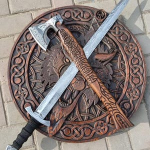 May include: A circular wooden shield featuring a carved wolf head and intricate knotwork patterns. A metal sword with an etched blade and a black wrapped handle is crossed with a wooden axe that has a metal head and carved handle.