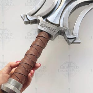 May include: A close-up of a silver-colored sword with a brown leather-wrapped handle. The sword has a decorative hilt with a stylized design and engraved text. The handle is held by a person.