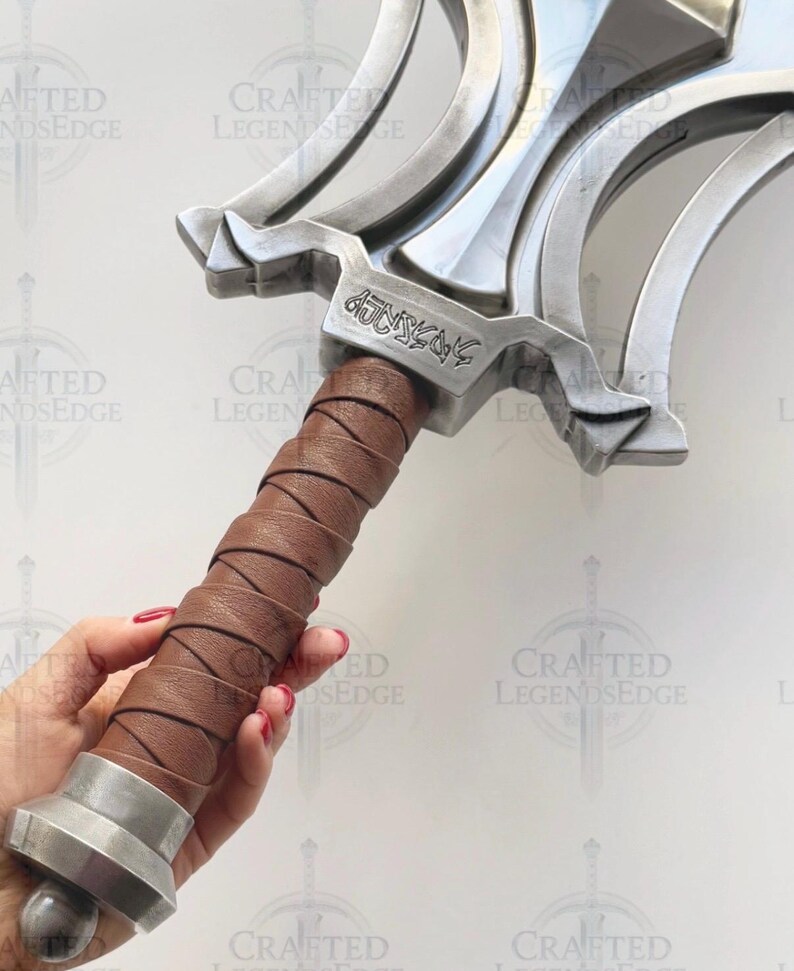 May include: A close-up of a silver-colored sword with a brown leather-wrapped handle. The sword has a decorative hilt with a stylized design and engraved text. The handle is held by a person.