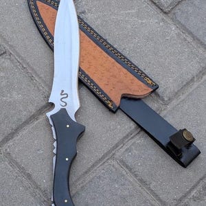 May include: A silver-bladed knife with a black handle and a brown leather sheath. The knife features a snake design near the blade's base. The sheath is brown with black accents and stitching. The knife and sheath are displayed on a brick surface.