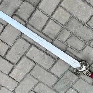 May include: A long, silver sword with a decorative handle. The handle features a burgundy grip and a gold-colored pommel. The sword's blade is straight and tapers to a sharp point. The sword is lying on a gray brick surface.