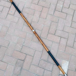 May include: A double-edged spear with a wooden shaft and two metal spearheads. The shaft is wrapped in black leather near the center and has a silver band. The spear is approximately 72 inches long.