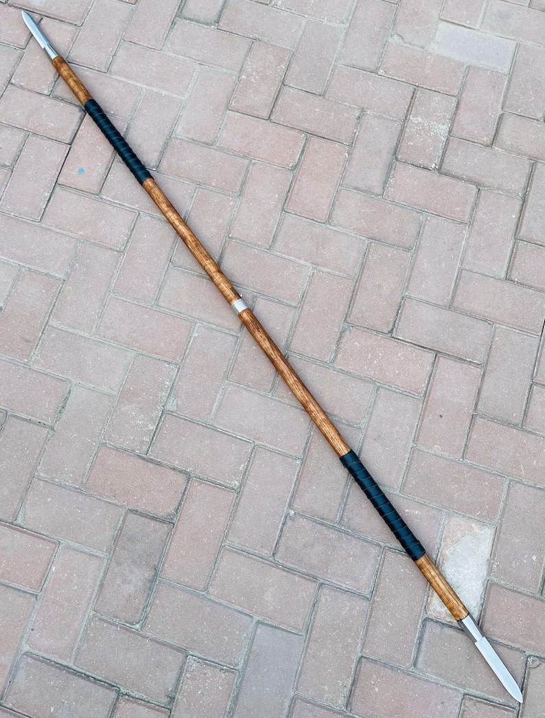 May include: A double-edged spear with a wooden shaft and two metal spearheads. The shaft is wrapped in black leather near the center and has a silver band. The spear is approximately 72 inches long.