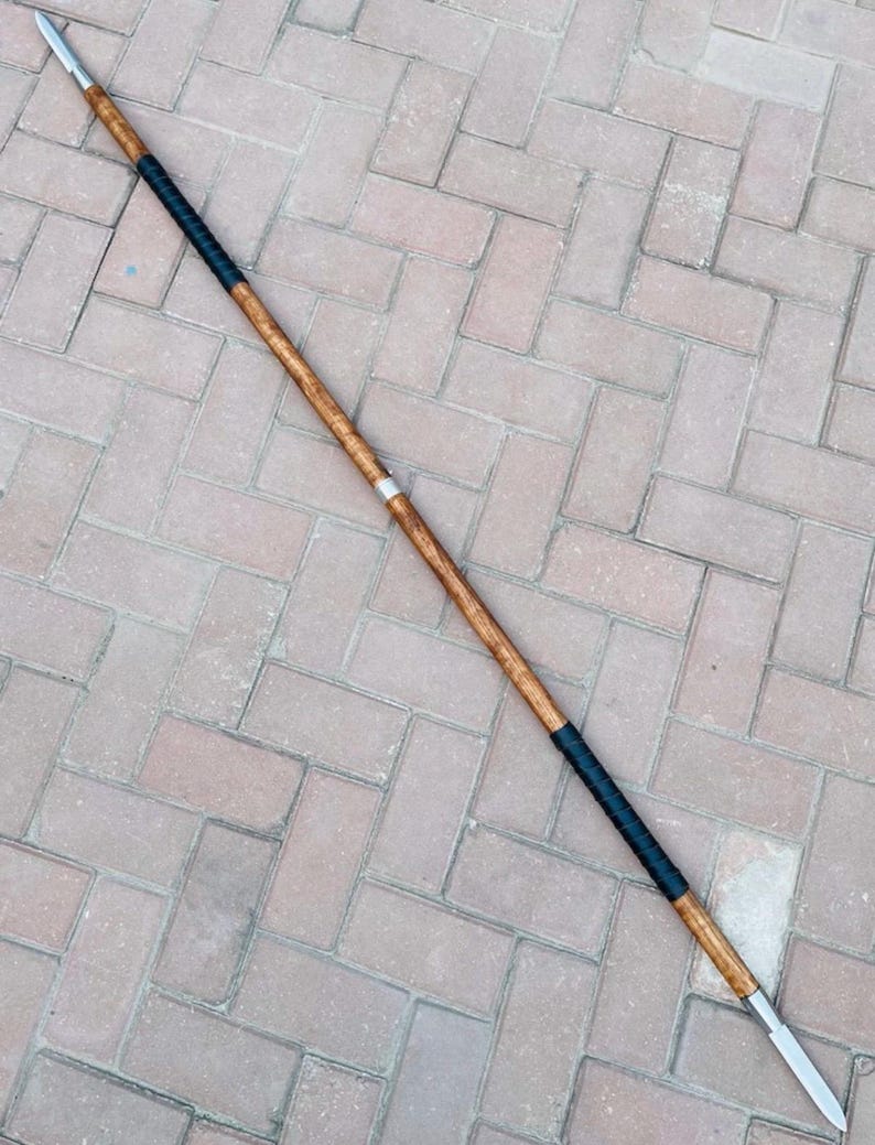 May include: A long spear with a wooden shaft, metal spearheads, and black wrapped grips. The spear has a light brown wooden shaft with a silver band near the center.
