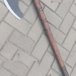 May include: A long-handled axe featuring a dark grey, curved metal blade with a sharpened silver edge. The handle is constructed from brown wood with a textured finish, wrapped in a dark brown leather strap in a crisscross pattern along the shaft.